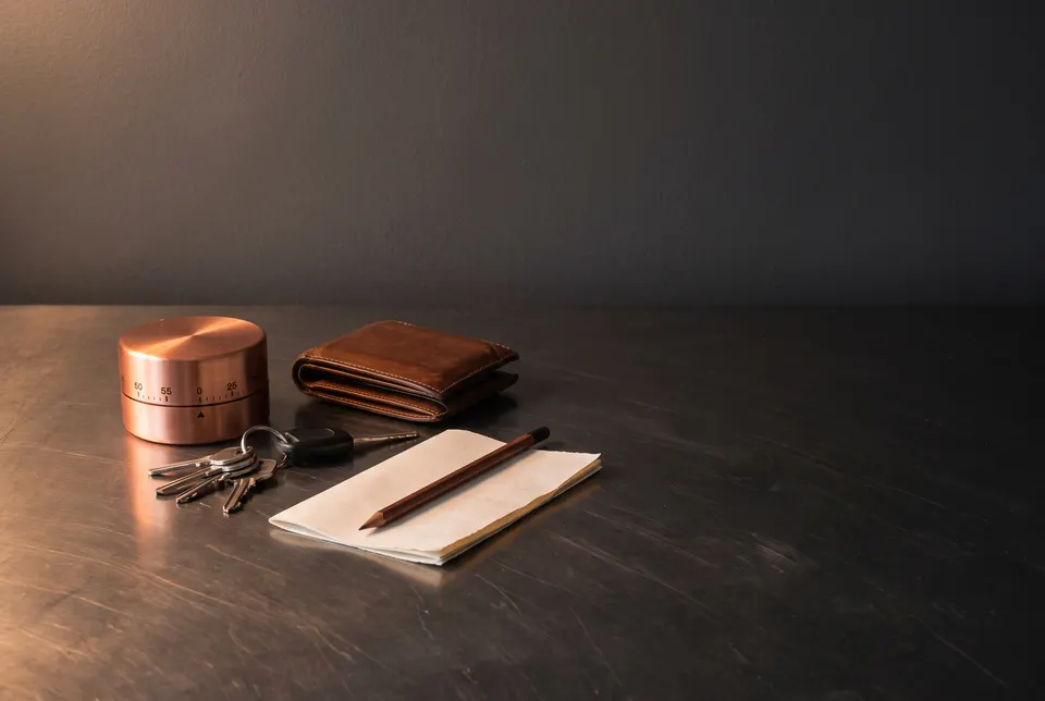 Generated scene: a copper kitchen timer, a closed leather wallet, car keys and a blank note with a pencil on a dark steel table.
